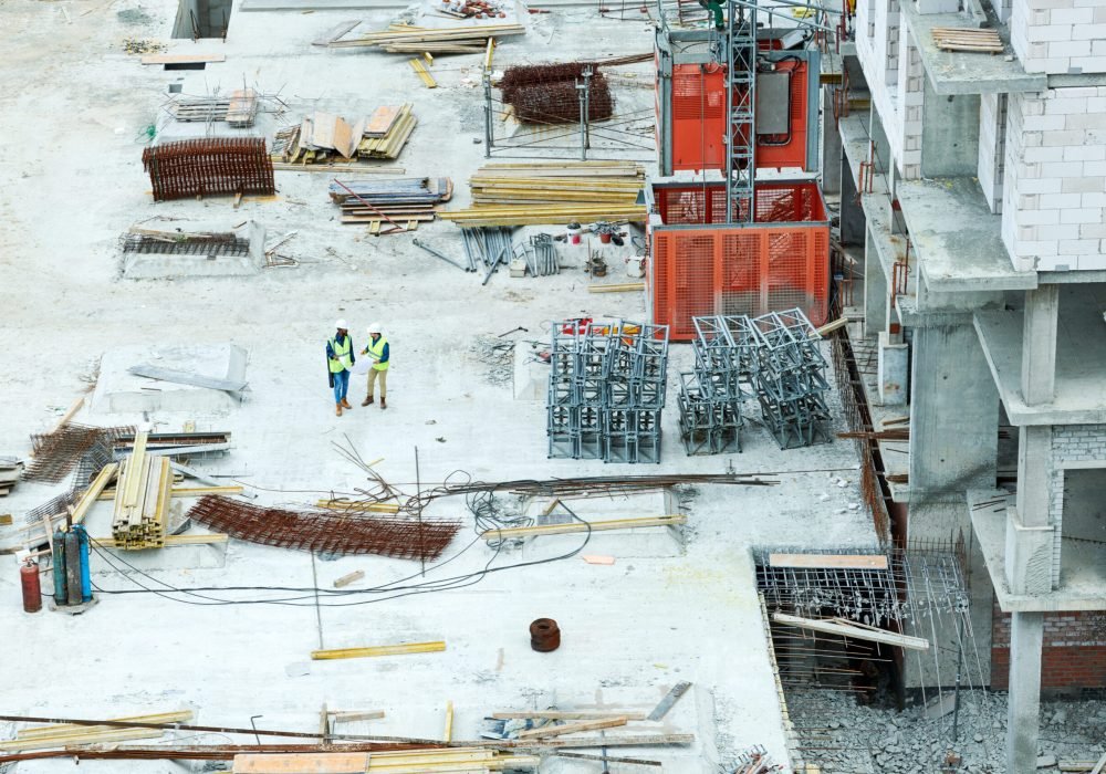 Directly above view of modern workmen in safety vests and hardhats standing on construction site with stacks of building materials and discussing plan